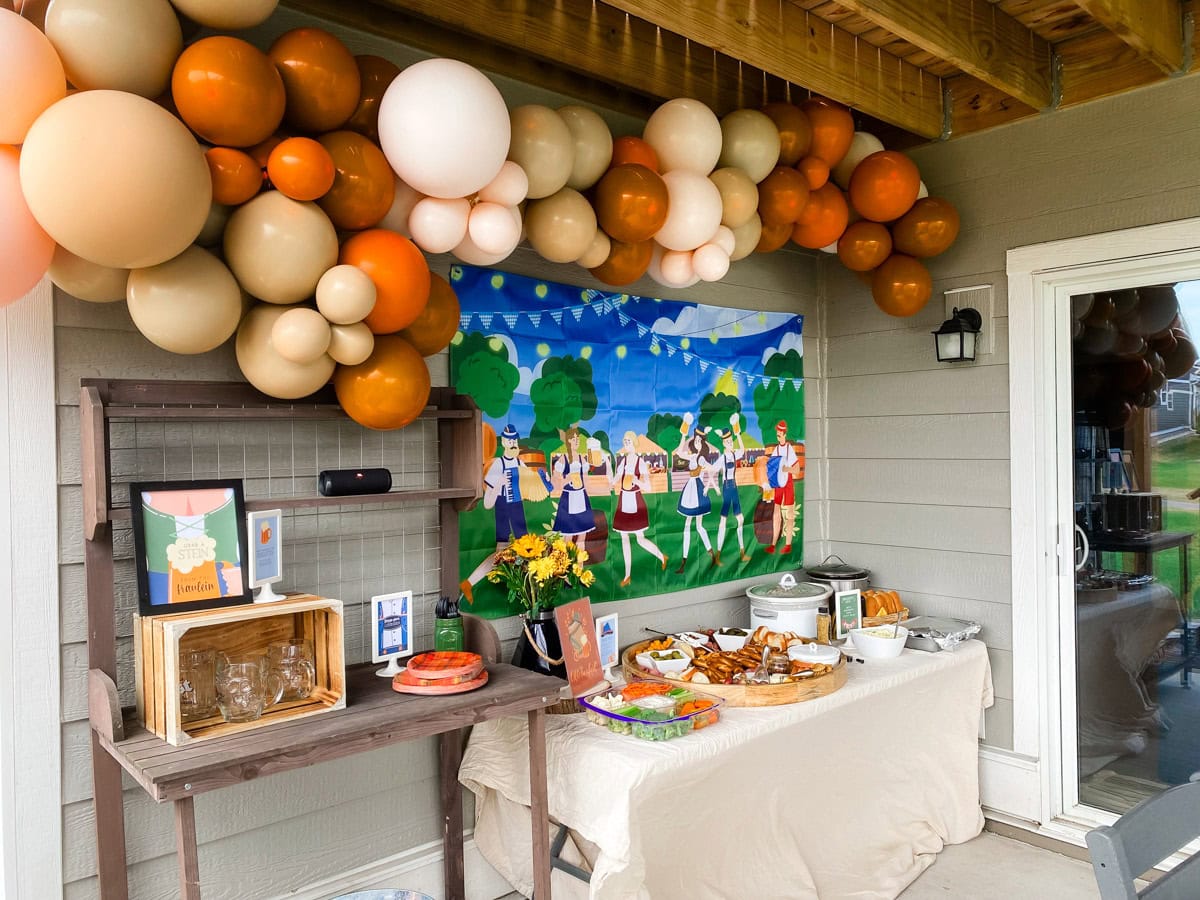 Adult Oktoberfest party fraulein theme backyard biergarten setup with orange and tan balloon garland, Oktoberfest backdrop, and food table