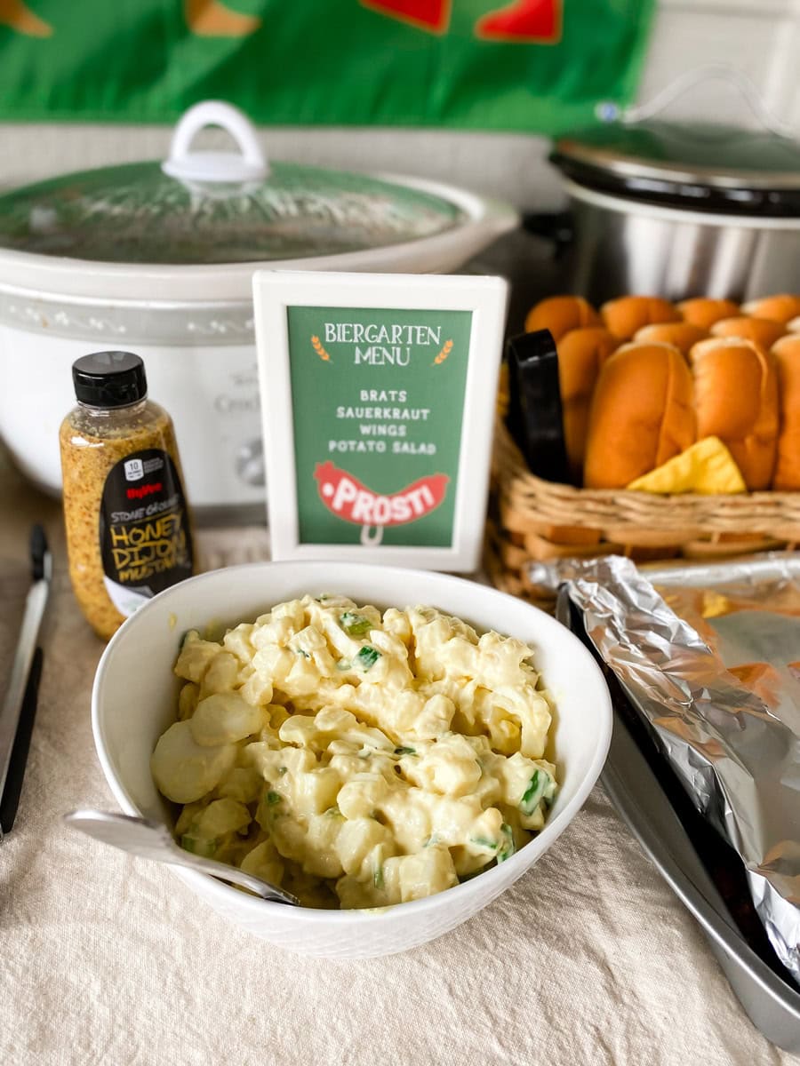 Biergarten Menu printable sign listing brats, sauerkraut, wings, and potato salad next to a bowl of potato salad at an adult Oktoberfest party