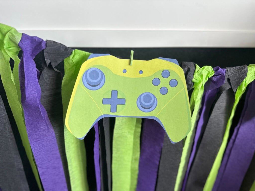 Lime green paper game controller cutout decoration pinned to purple, black, and lime green crepe paper streamer backdrop for gamer birthday party