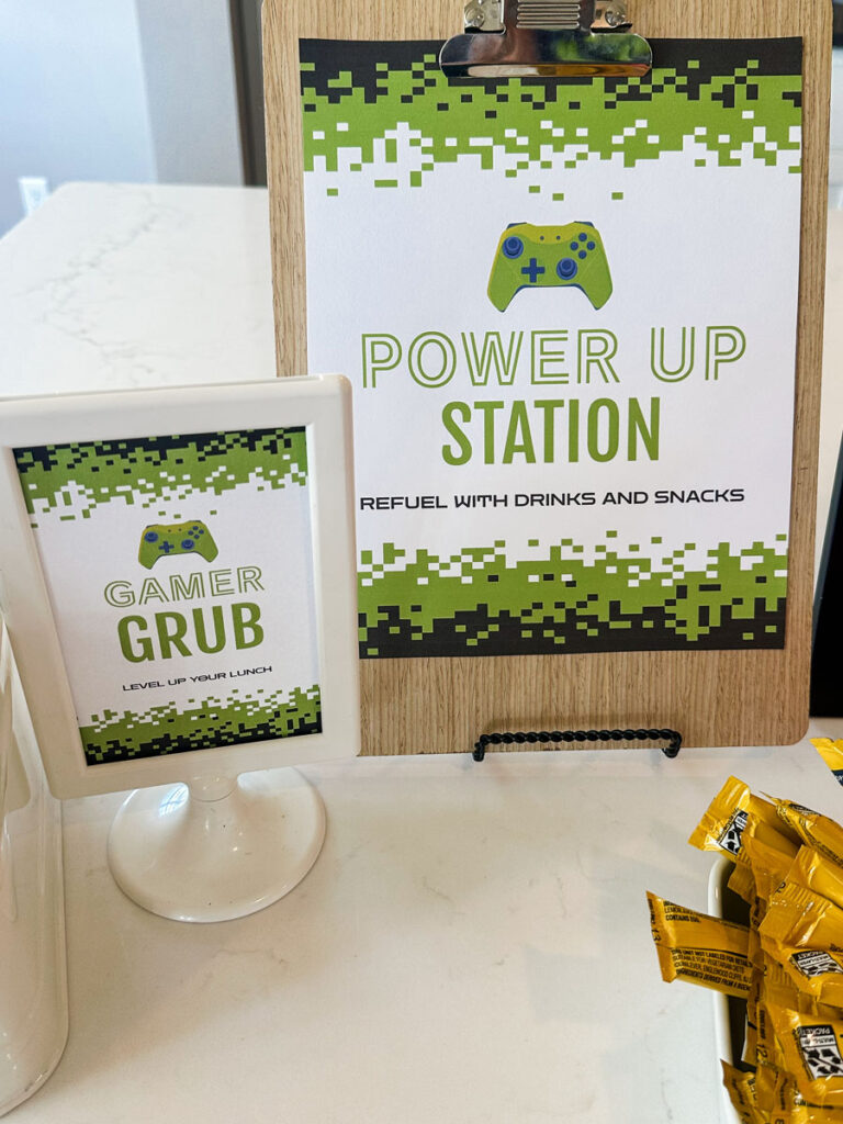 Gamer Grub — Level Up Your Lunch" small framed printable sign and "Power Up Station — Refuel with Drinks and Snacks" clipboard sign at gamer birthday party food station
