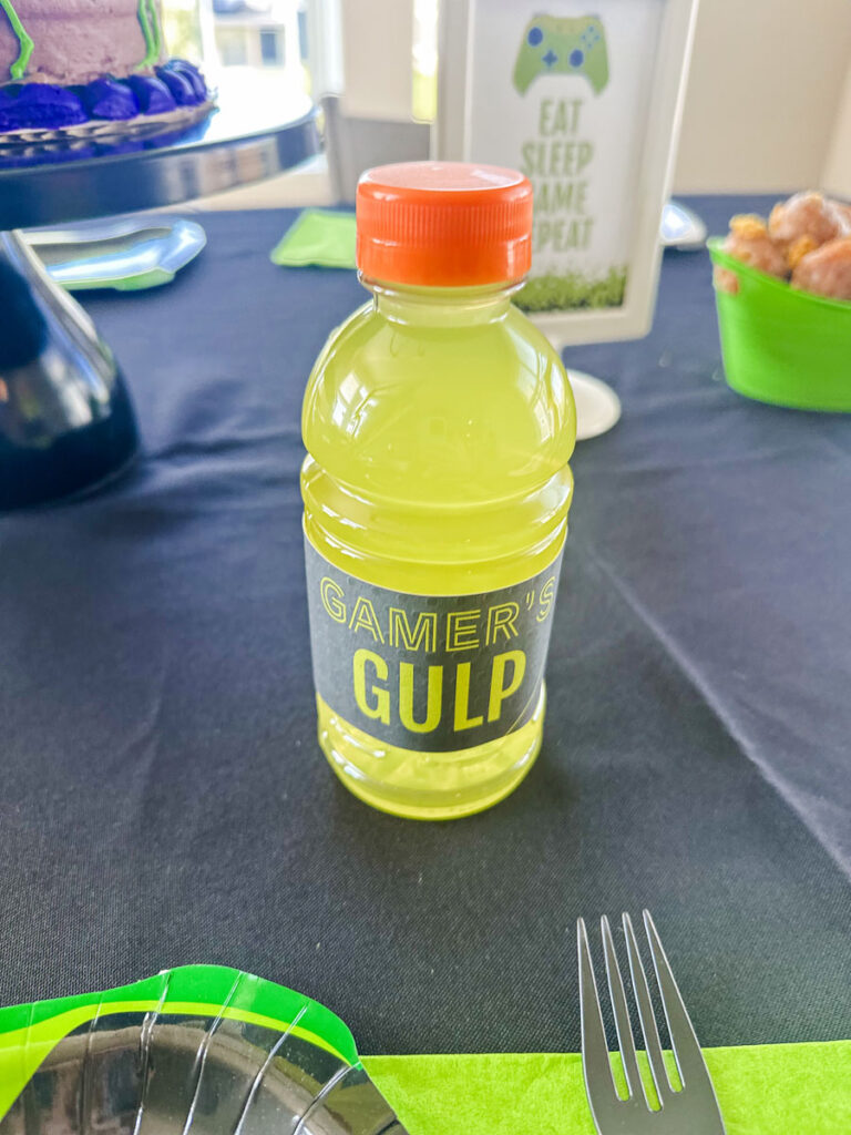 "Gamer's Gulp" custom-labeled Gatorade bottle at gamer birthday party dining table with Eat Sleep Game Repeat sign in background