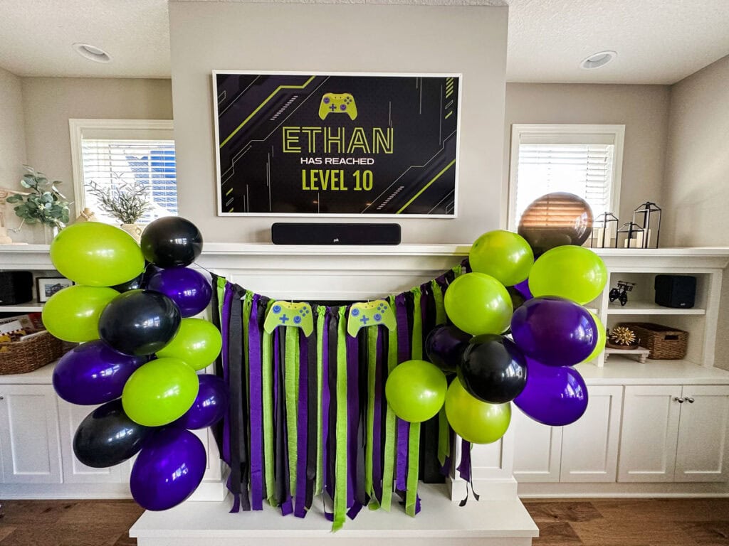 Gamer birthday party backdrop on living room fireplace mantel with lime green, black, and purple crepe paper streamers, paper game controller cutouts, balloon clusters, and TV displaying "Ethan Has Reached Level 10" custom sign