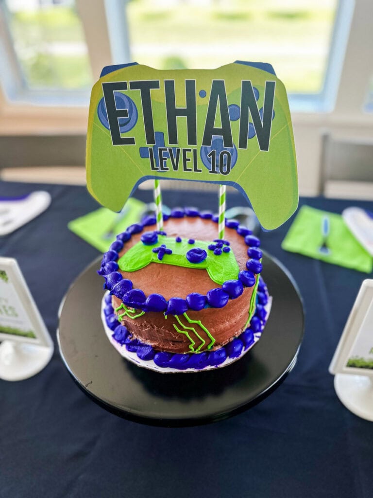 Gamer birthday cake with blue frosted border, lime green game controller pipe design, and "Ethan Level 10" game controller-shaped printable cake topper on black cake stand