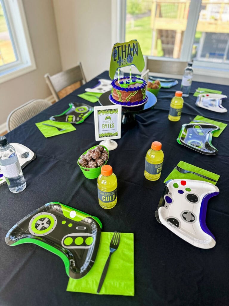 Gamer birthday party dining table with black tablecloth, game controller-shaped paper plates, lime green napkins, Gamer's Gulp Gatorade bottles, birthday cake, and Tasty Bytes printable sign