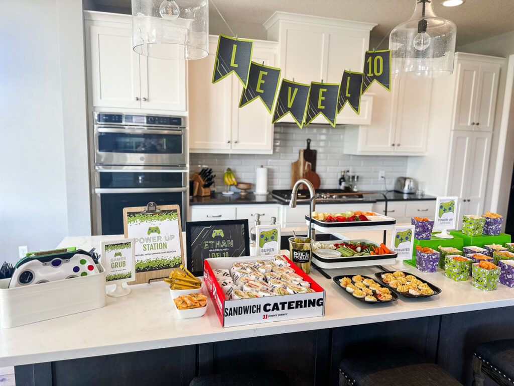 Gamer birthday party Power Up Station food buffet on kitchen island with Level 10 banner, Jimmy John's catering, fruit and veggie tiered platter, and individual gaming-print chip boxes