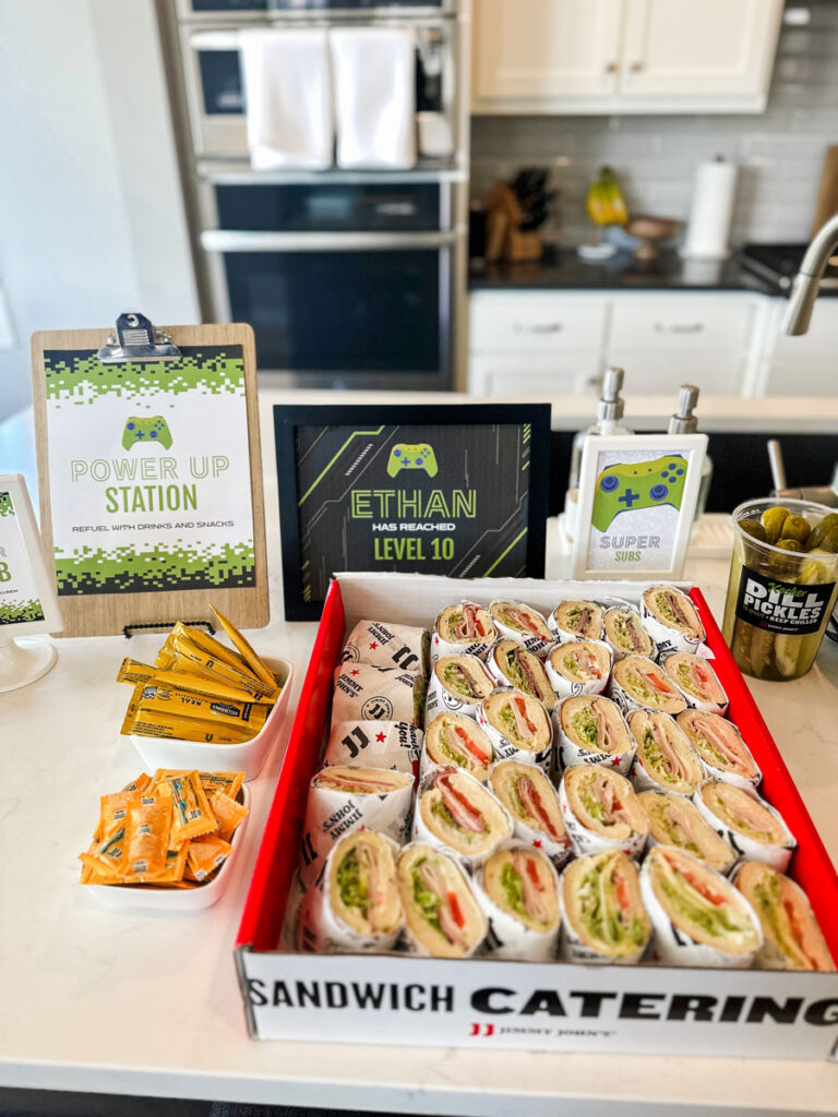 Jimmy John's sandwich catering box labeled "Super Subs" at gamer birthday party food station with Power Up Station clipboard sign, Ethan Has Reached Level 10 black framed sign, and dill pickles
