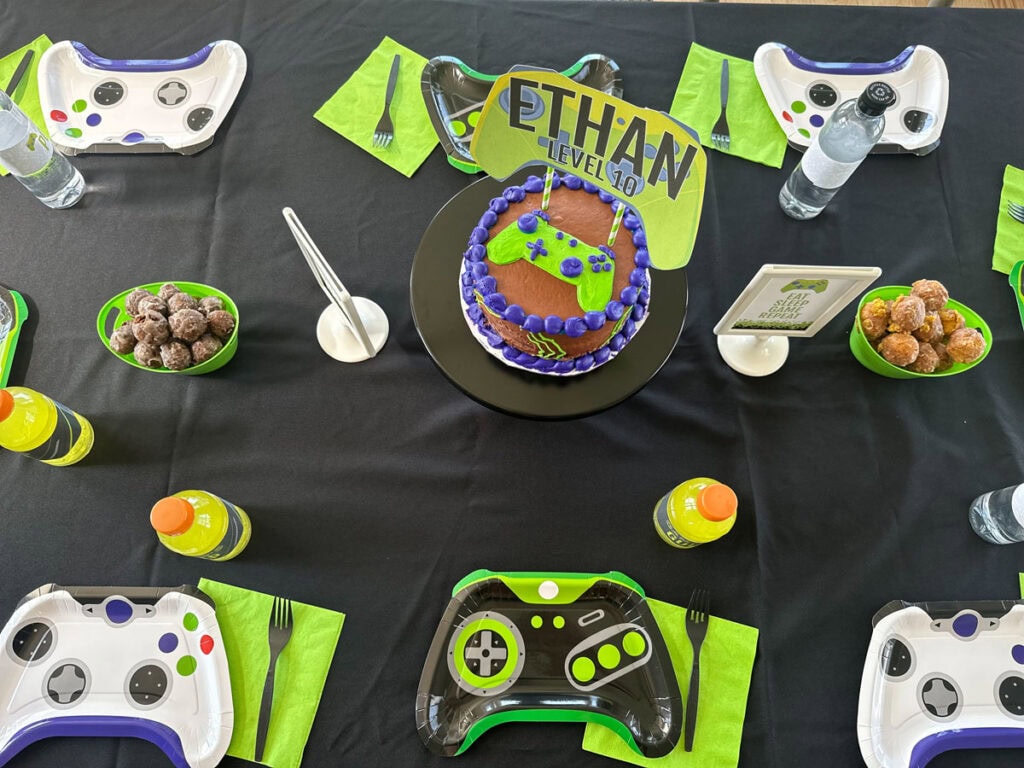 Overhead view of gamer birthday party dining table with controller plates, birthday cake with Level 10 cake topper, chocolate bites in lime green bowls, and Eat Sleep Game Repeat printable sign