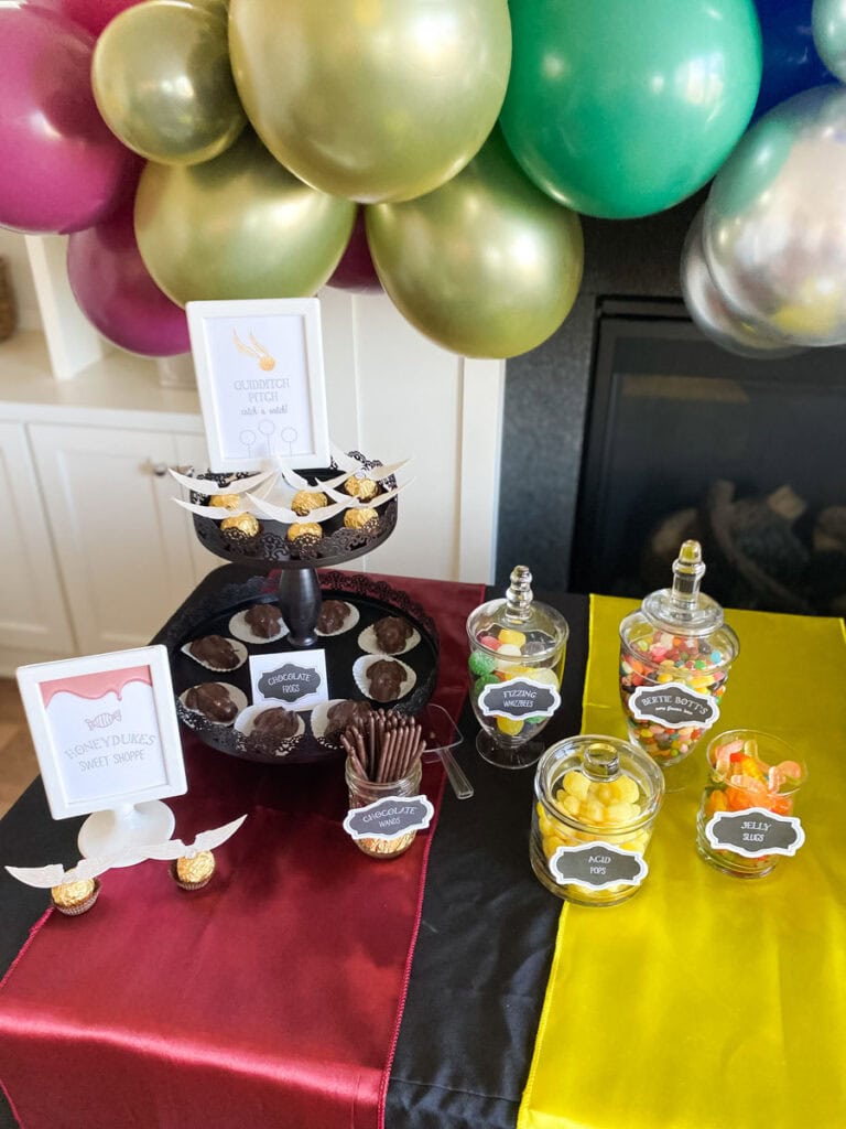 Harry Potter Honeydukes Sweet Shoppe dessert table with printable candy jar labels for Acid Pops, Fizzing Whizzbees, Chocolate Wands, and Jelly Slugs on a black tablecloth with Hogwarts house color table runners