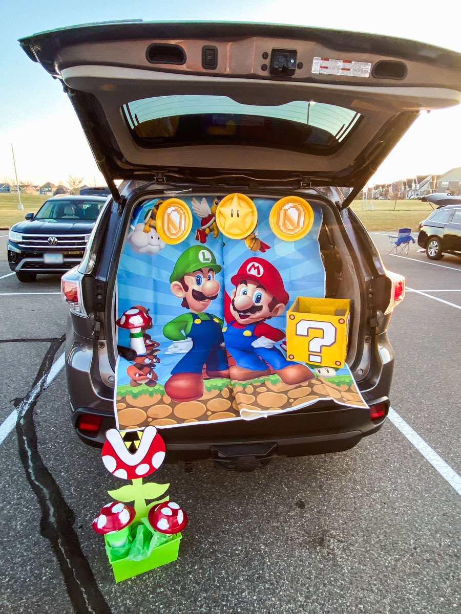 Mario trunk or treat SUV with Mario and Luigi backdrop, paper plate coins and star, yellow question mark box, and DIY piranha plant with red mushrooms
