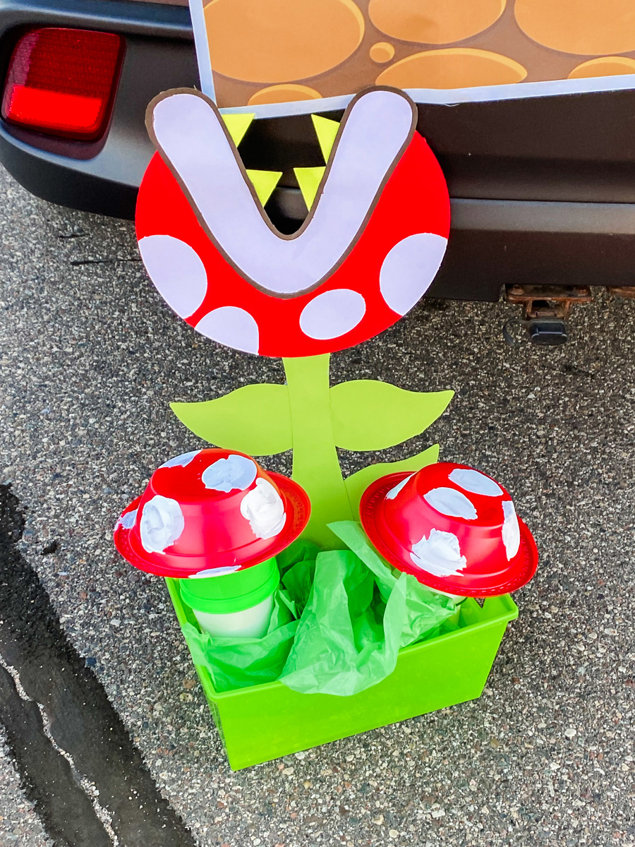 DIY piranha plant for a Mario trunk or treat with red and white spotted head, green stem and leaves, and two red mushroom cups in a green bin with green tissue paper