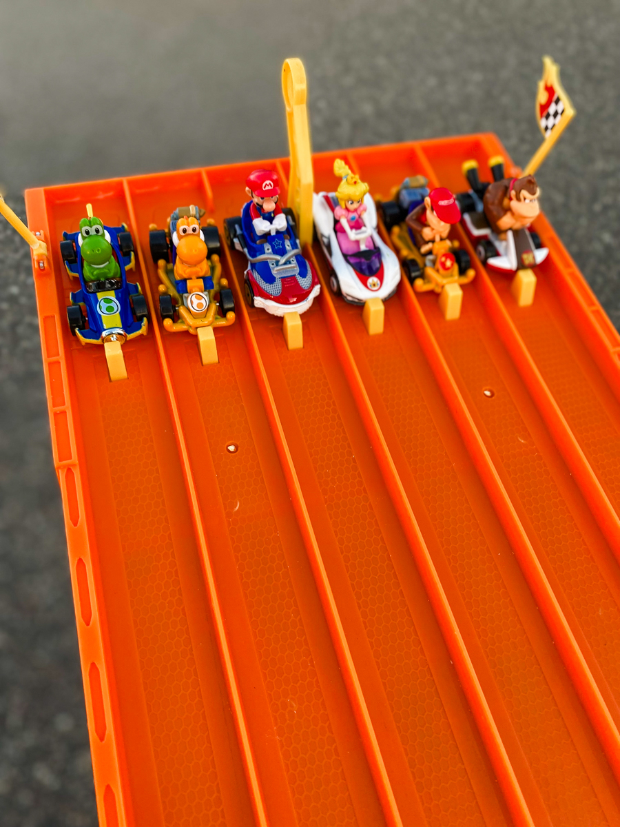 Hot Wheels Mario Kart cars including Yoshi, Mario, Princess Peach, and Donkey Kong lined up on an orange six-lane race track for a trunk or treat game