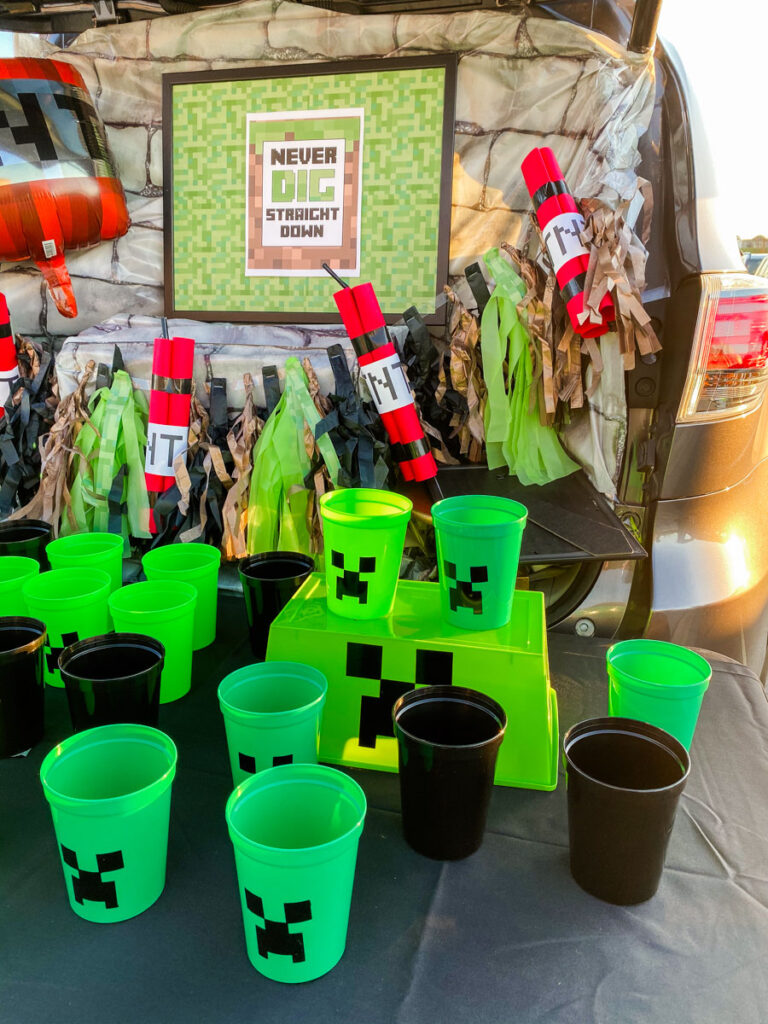 Daytime view of Minecraft trunk or treat trunk display with stone backdrop, TNT garland, TNT foil balloon, and rows of green and black Creeper cups