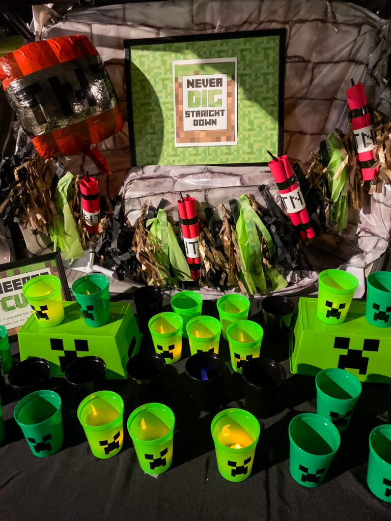 Minecraft trunk or treat backdrop at dusk with stone table cover, framed Never Dig Straight Down printable sign, TNT dynamite garland, and glowing green Creeper Cup game