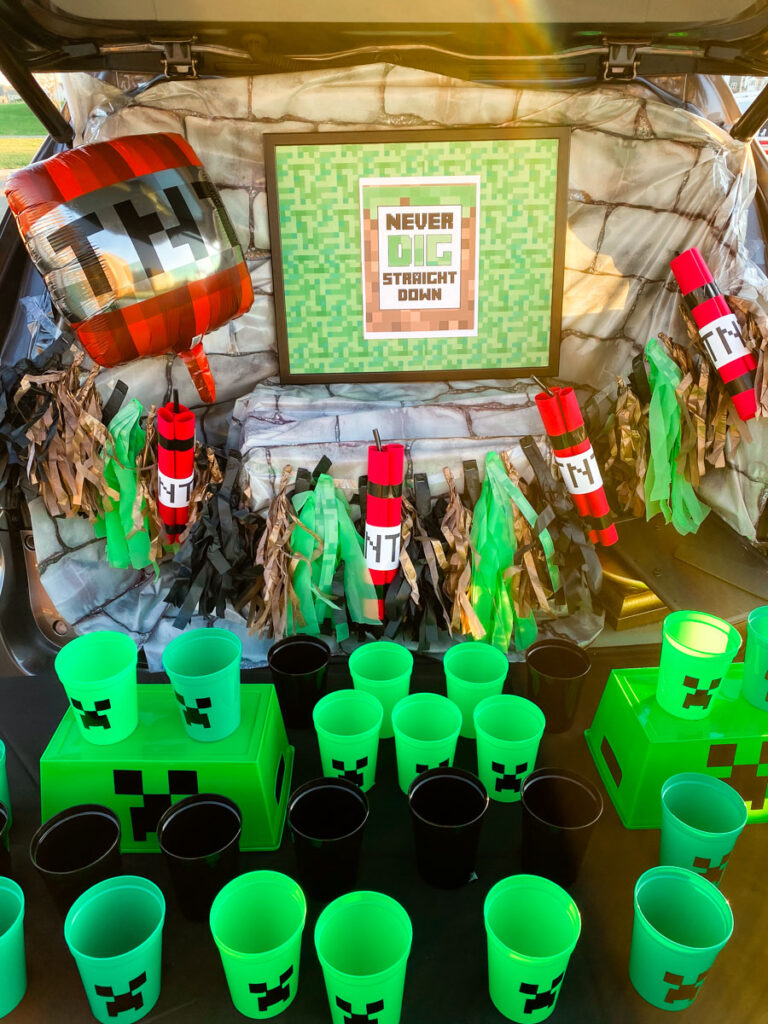 Minecraft trunk or treat setup with stone backdrop, TNT dynamite garland, TNT balloon, and green Creeper cups lined up in a car trunk