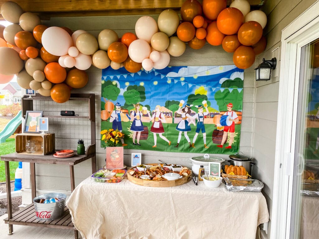 Adult Oktoberfest party fraulein theme backyard biergarten setup with orange, tan, and blush balloon garland, Oktoberfest backdrop with dancers, and food table