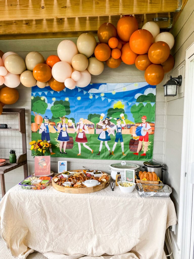 oktoberfest party backdrop with dancing people in dirndls and lederhosen, balloon garland, and table with oktoberfest party food