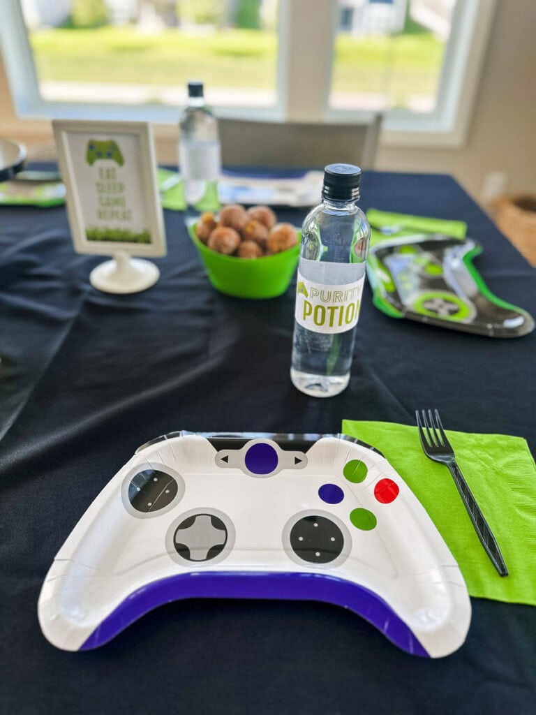 "Purity Potion" labeled water bottle and game controller-shaped paper plate at gamer birthday party dining table with Eat Sleep Game Repeat sign in background