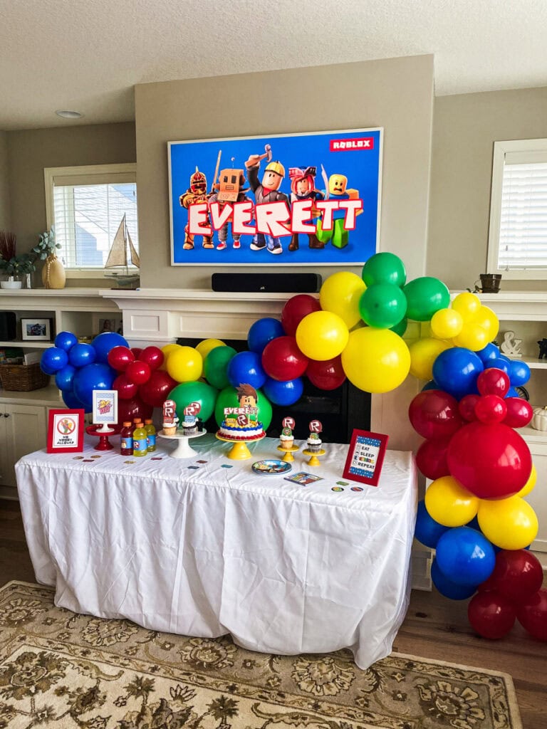 Wide view of Roblox birthday party room with balloon garland arch, dessert table, birthday cake, cupcakes, and printable signs
