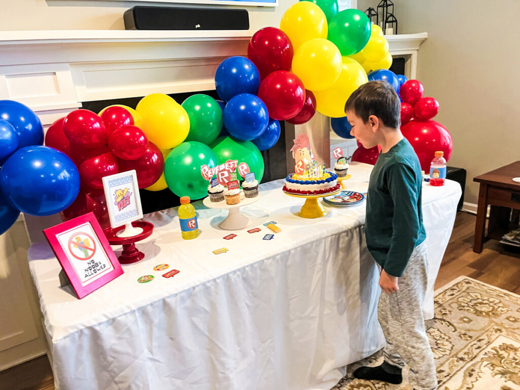 Birthday boy standing at his Roblox birthday party dessert table looking at the cake and balloon garland arch