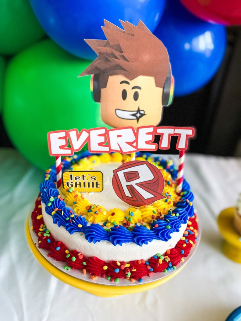 Roblox birthday party cake on yellow cake stand with red and blue buttercream frosting, rainbow sprinkles, Roblox R logo, and personalized cake topper