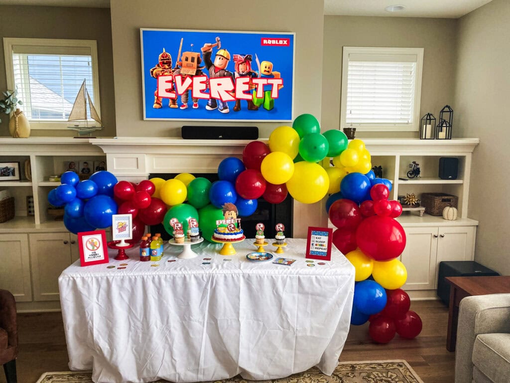 Roblox birthday party full setup with large red, blue, yellow, and green balloon garland arch over dessert table and custom Everett TV display