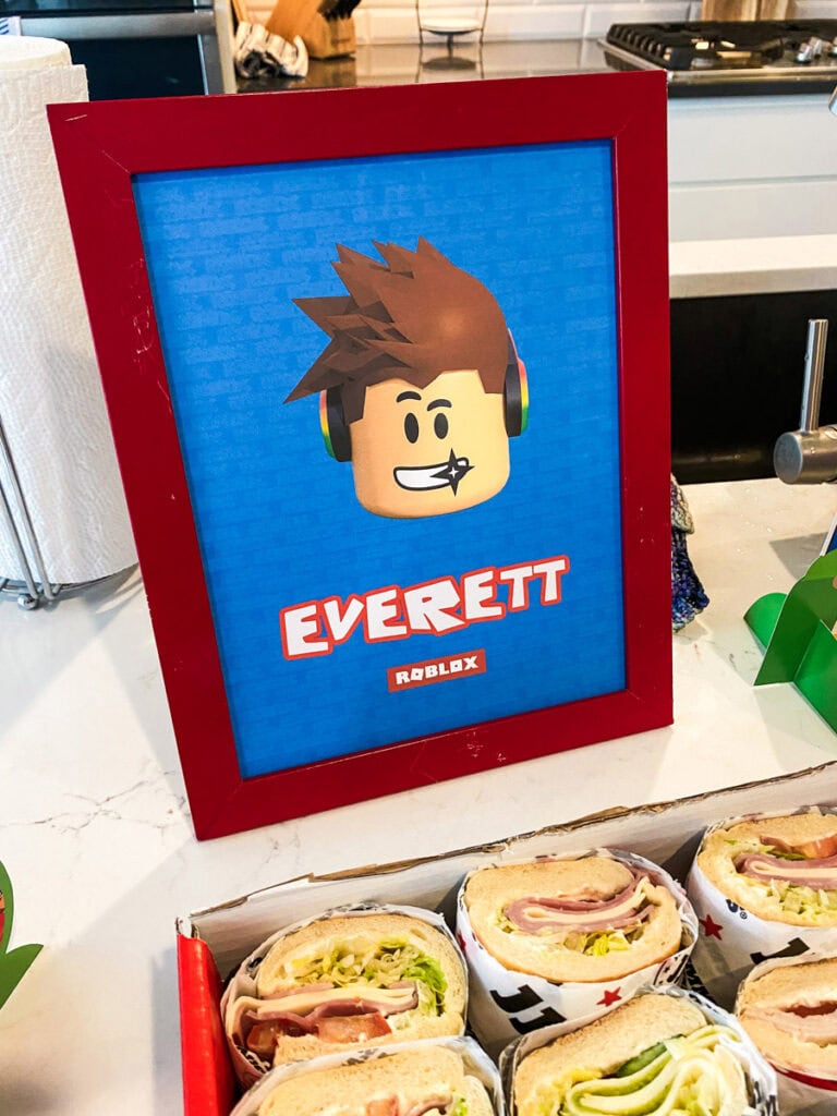 Custom Roblox printable portrait sign in red frame beside Jimmy John's sandwich catering box at Roblox birthday party food station