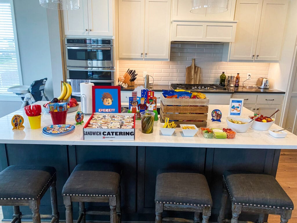 Roblox birthday party food spread on kitchen island with Jimmy John's sandwich catering, custom Roblox decorations, salad, fruit, and snacks