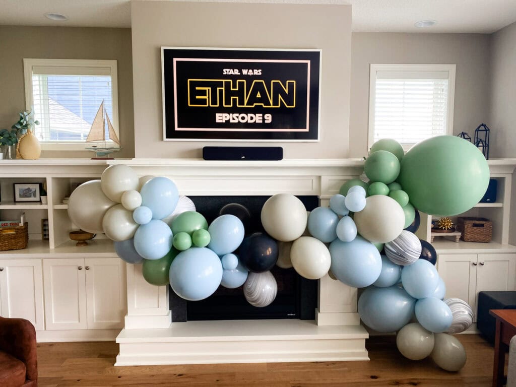 Muted organic balloon garland in dusty blue, sage green, cream, and black above fireplace with Star Wars TV display