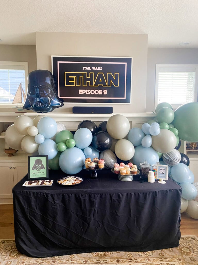 Full view of muted Star Wars birthday party backdrop with balloon garland, TV display, and dessert table