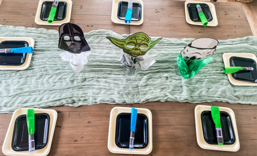 Star Wars birthday party dining table with sage green cheesecloth runner, character mask centerpieces, black plates, and lightsaber napkin rings